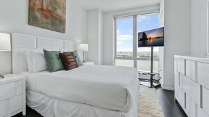 Ein modernes Schlafzimmer mit einem großen Bett, weißen Nachttischen, einem an der Wand montierten Fernseher und Blick auf die Stadt durch große Fenster.