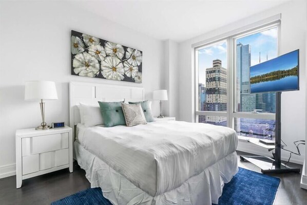 A modern bedroom with a large bed, white nightstands, a city view, and a painting above the bed. - High-Rise NYC, Furnished 1BR w. Views & Full Kitchen (New York)