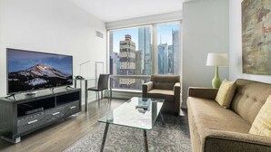 Un moderno salón con una televisión grande, una mesa de centro de cristal, un sofá y vistas a edificios de la ciudad a través de amplias ventanas.