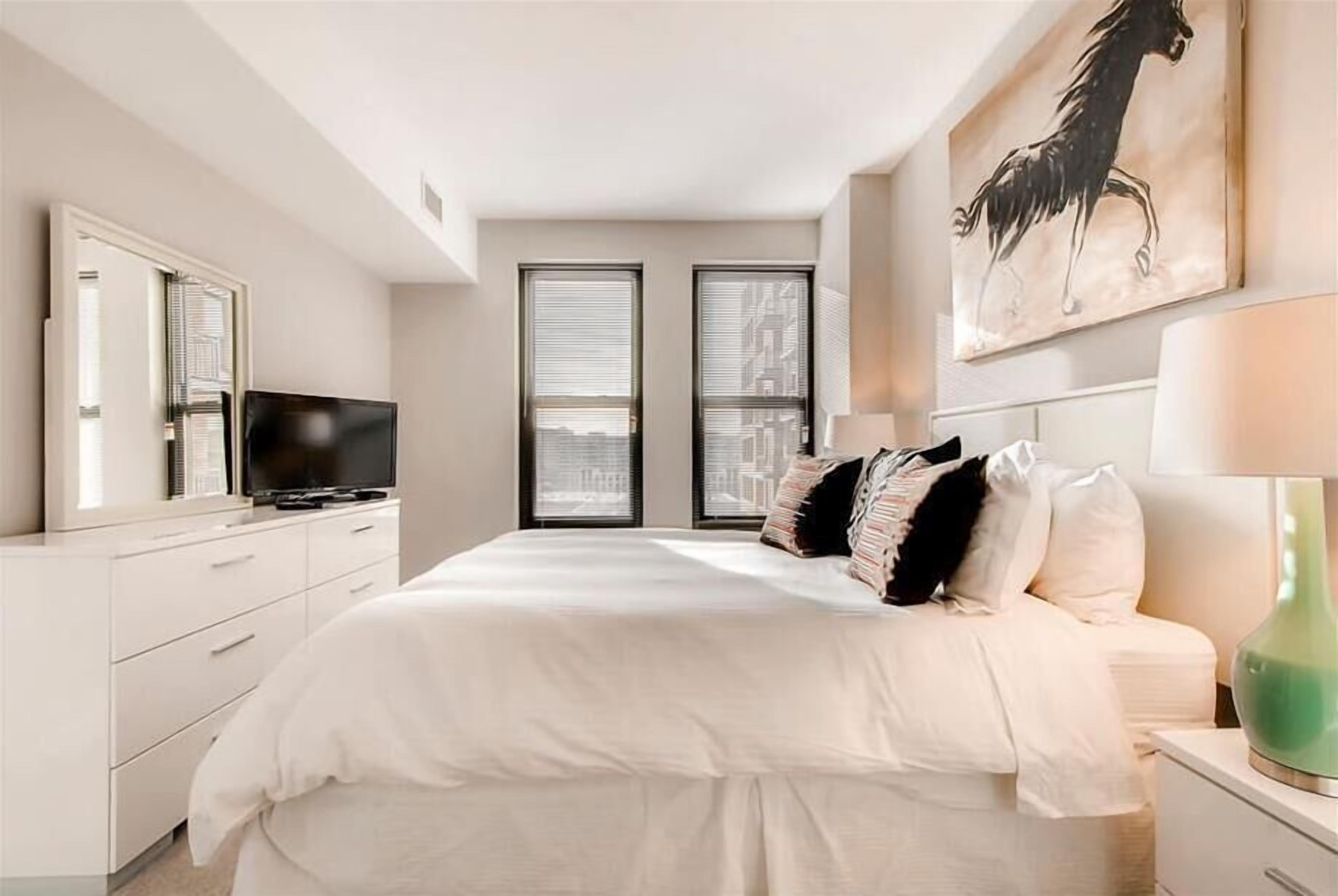 A modern bedroom with a large bed, a television, and a city view through the windows.