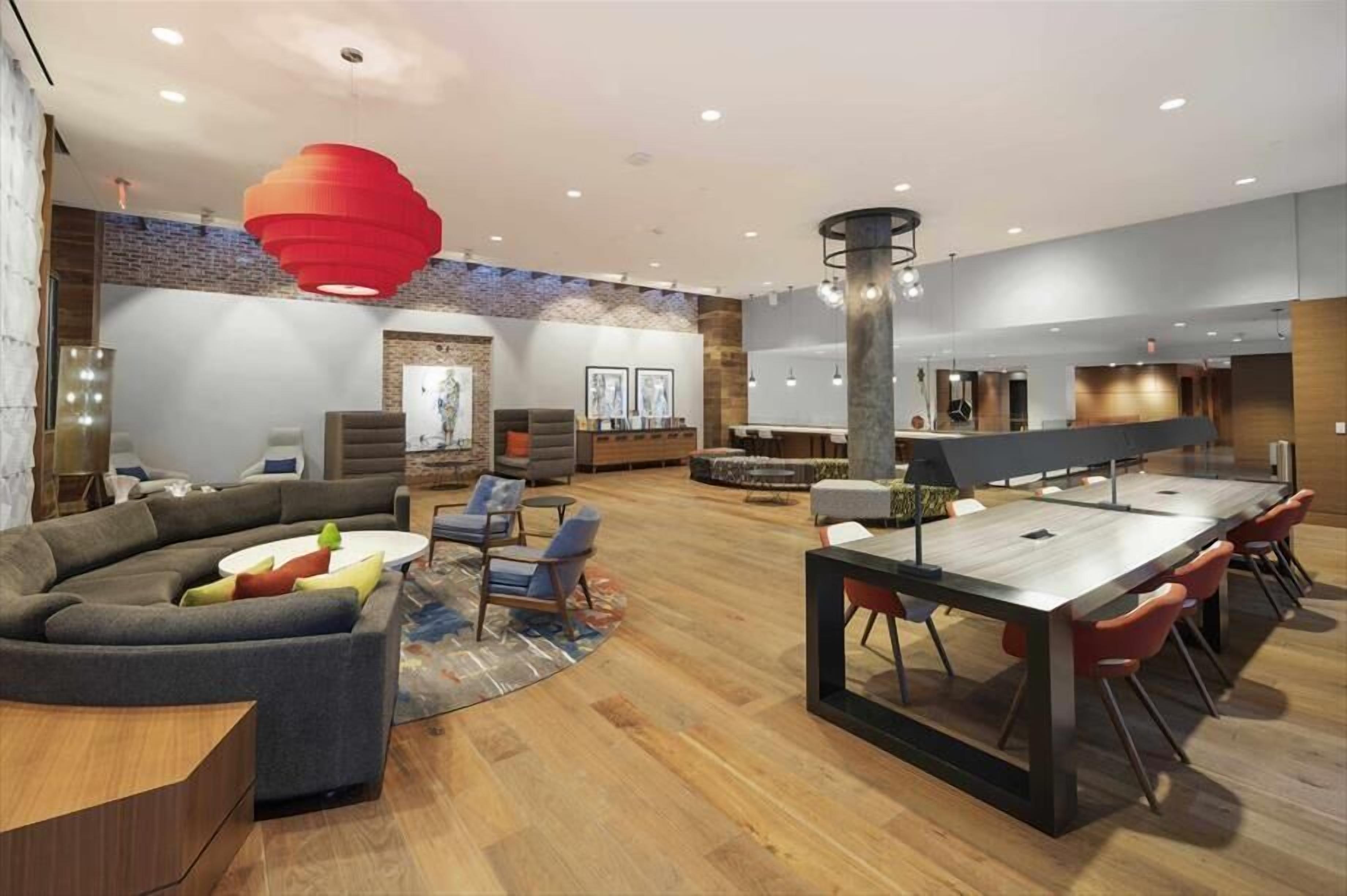 Een moderne hotel lobby met een gebogen bank, een ronde salontafel, een eetgedeelte met een lange tafel en rode stoelen, en een grote rode hanglamp.