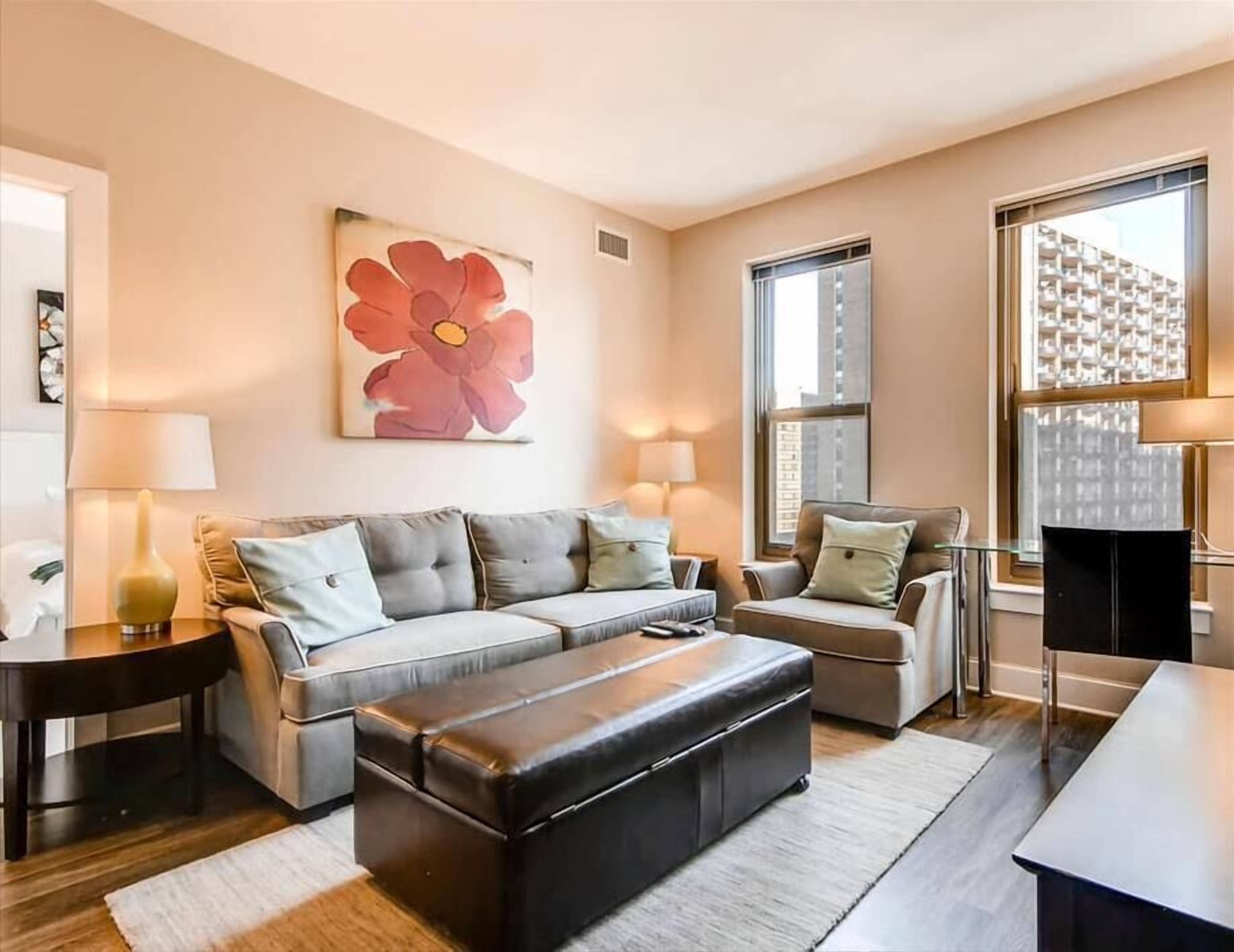 Ein modernes Wohnzimmer mit einem grauen Sofa, einem schwarzen Hocker und einem großen Fenster mit Blick auf die Stadt.