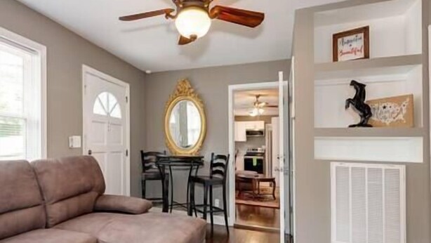 A living room with a brown sofa, a dining area with a table and chairs, a ceiling fan, and a hallway leading to a kitchen. - Downtown Winston's - 2 BR - Queens - Pad 2 (Winston-Salem)