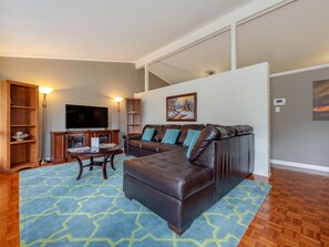 A modern living room with a sectional sofa, a flat-screen TV, wooden shelving, and a patterned rug. - Mid-Century Triad Gem: 3 Beds, 2 Baths (Winston-Salem)