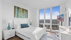 A modern bedroom with a large bed, a cityscape view through a large window, and a painting on the wall.