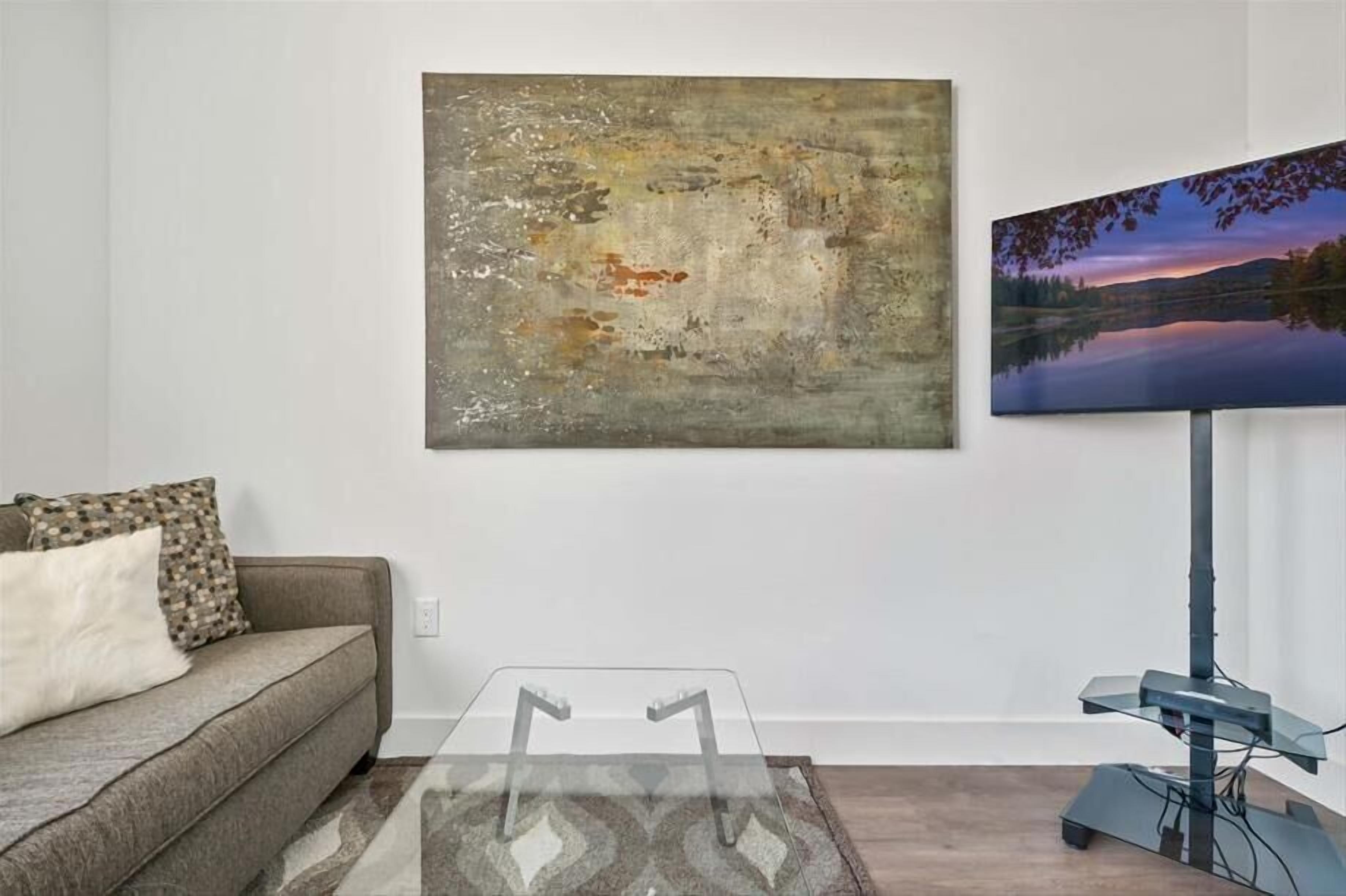 Un salon avec un canapé gris, une table basse en verre, un grand tableau abstrait et un téléviseur à écran plat posé sur un meuble.