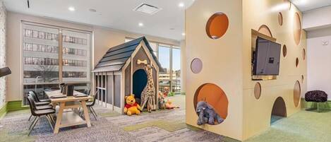 A children's play area with a giraffe-themed playhouse, a table with chairs, and a television mounted on a wall.