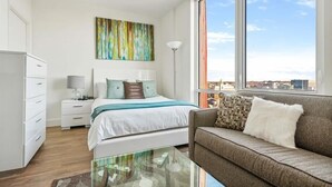 A modern bedroom with a bed, a sofa, a glass coffee table, and a large window with a city view.