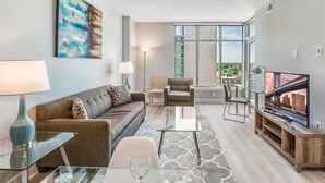 Un salon moderne avec un canapé, une table basse et un téléviseur. Une grande fenêtre offre une vue sur la ville.