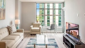 A modern living room with a sofa, armchair, and TV, large windows with a city view, and a floor lamp.
