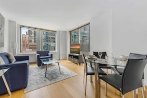 A modern living room with a sofa, a glass coffee table, a dining table with chairs, and a TV. Large windows offer views of buildings outside.