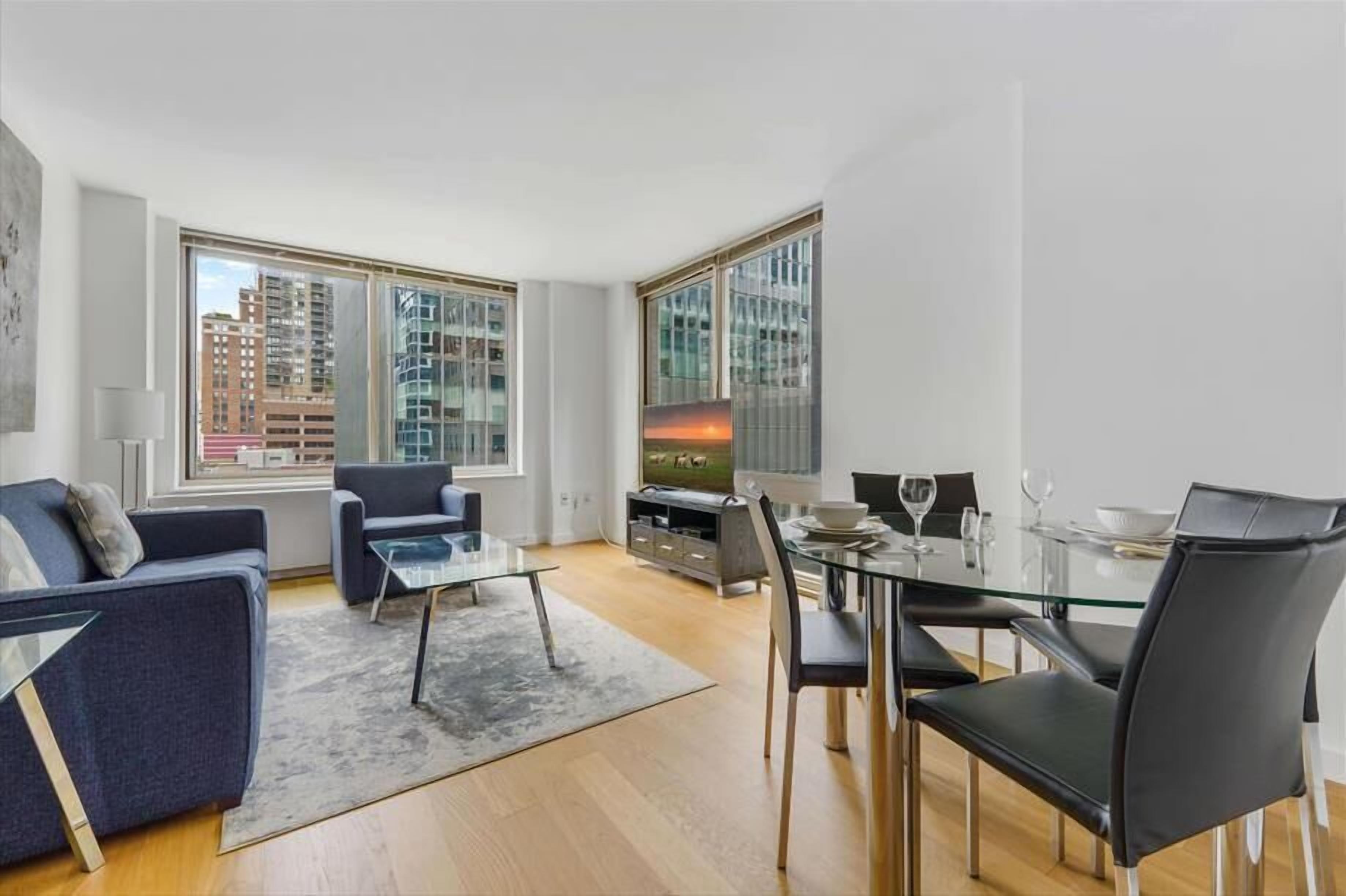 A modern living room with a sofa, a glass coffee table, a dining table with chairs, and a TV. Large windows offer views of buildings outside.