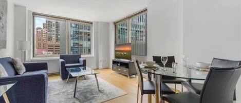 A modern living room with a sofa, a glass coffee table, a dining table with chairs, and a TV. Large windows offer views of buildings outside.
