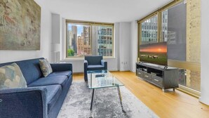 Una sala de estar moderna con un sofá, una mesa de centro de vidrio, un televisor sobre un mueble bajo y un ventanal con vista a la ciudad.