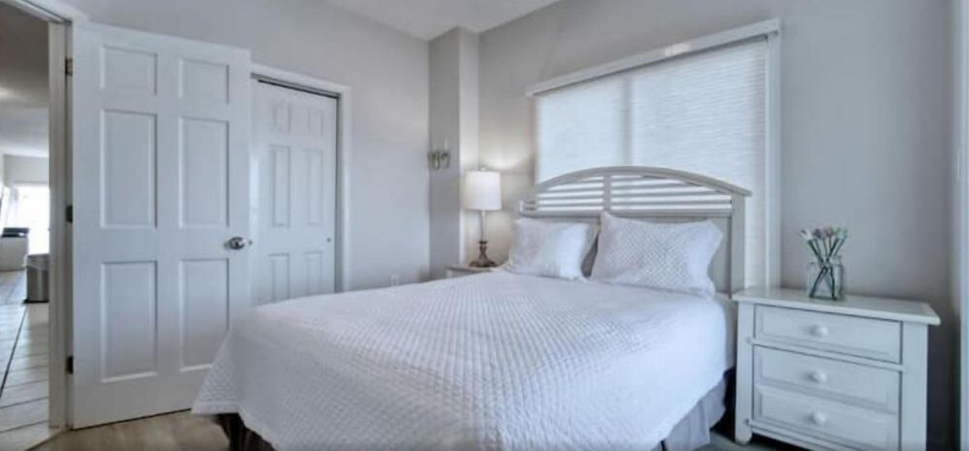 Une chambre à coucher avec un lit blanc, une table de chevet avec des tiroirs, une lampe et une porte.