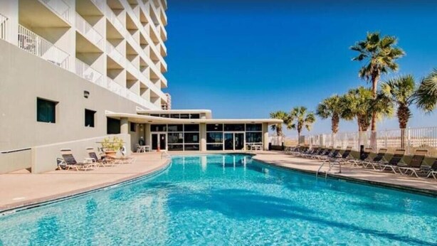 A swimming pool with lounge chairs and palm trees, surrounded by a building with a geometric facade. - 3 Bedrooms and 3 baths : Beach Facing Condo (Orange Beach)