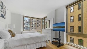 Un dormitorio moderno con una cama grande, un televisor y vistas a un paisaje urbano a través de amplios ventanales.