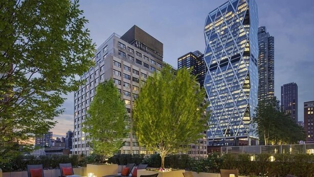 An outdoor seating area with lounge chairs and tables, surrounded by greenery and modern buildings in the background. - 54th Street 1 BR | Gym + Lounge | 24hr Concierge (New York)