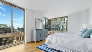 A modern bedroom with a large bed, a grey dresser, a TV, and a city view through large windows.