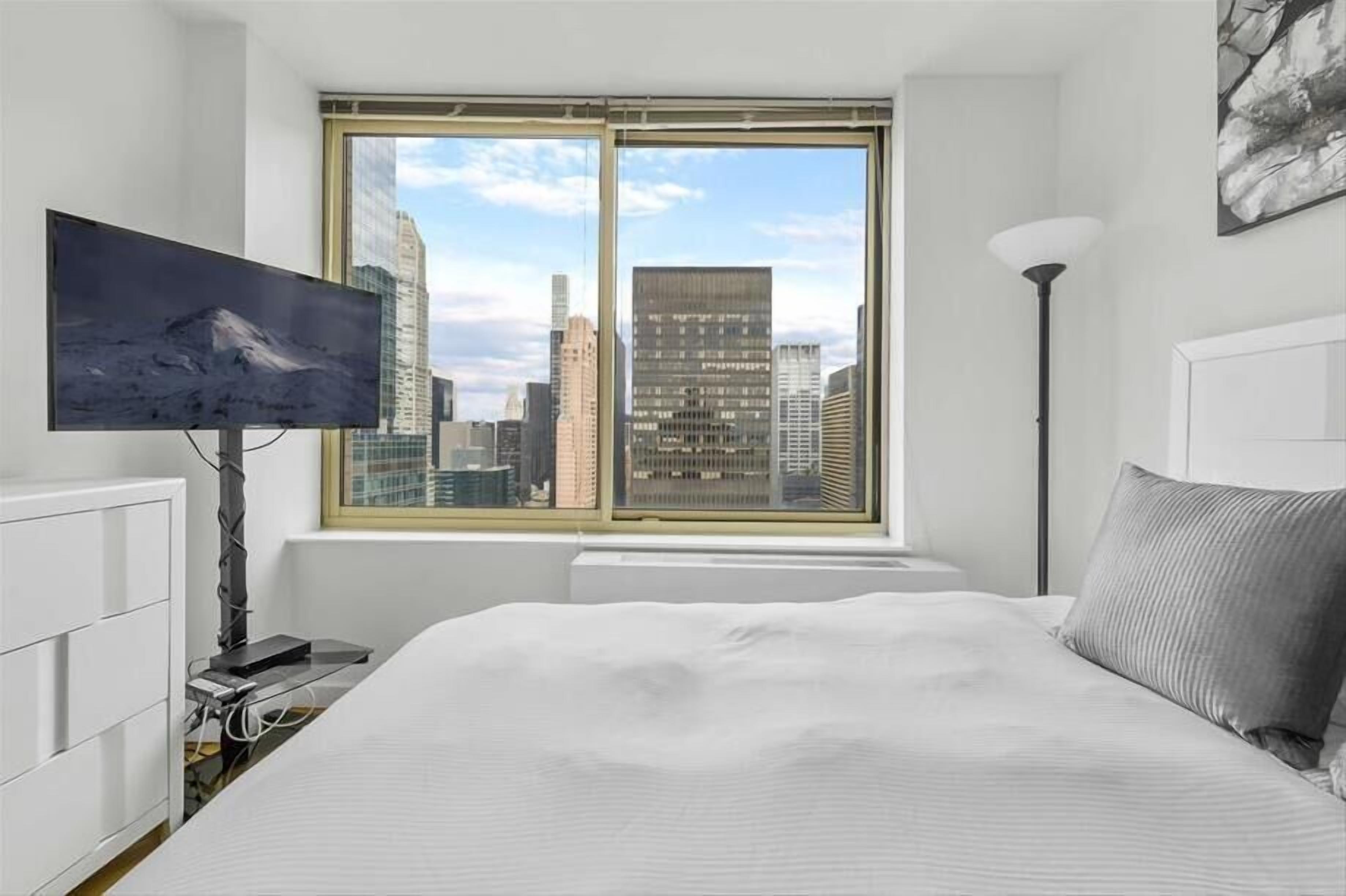 Un dormitorio con una cama grande, un televisor de pantalla plana en la pared, una ventana con vista a la ciudad y una lámpara de pie.