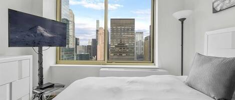 Un dormitorio con una cama grande, un televisor de pantalla plana en la pared, una ventana con vista a la ciudad y una lámpara de pie.