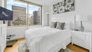 Une chambre moderne avec une grande fenêtre, un lit recouvert d’une literie blanche, un tableau représentant des fleurs et une table de chevet avec une lampe.