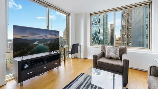 Un salon moderne doté d’un grand téléviseur à écran plat, d’un canapé, d’une table basse en verre et offrant une vue sur le paysage urbain grâce à de grandes fenêtres.