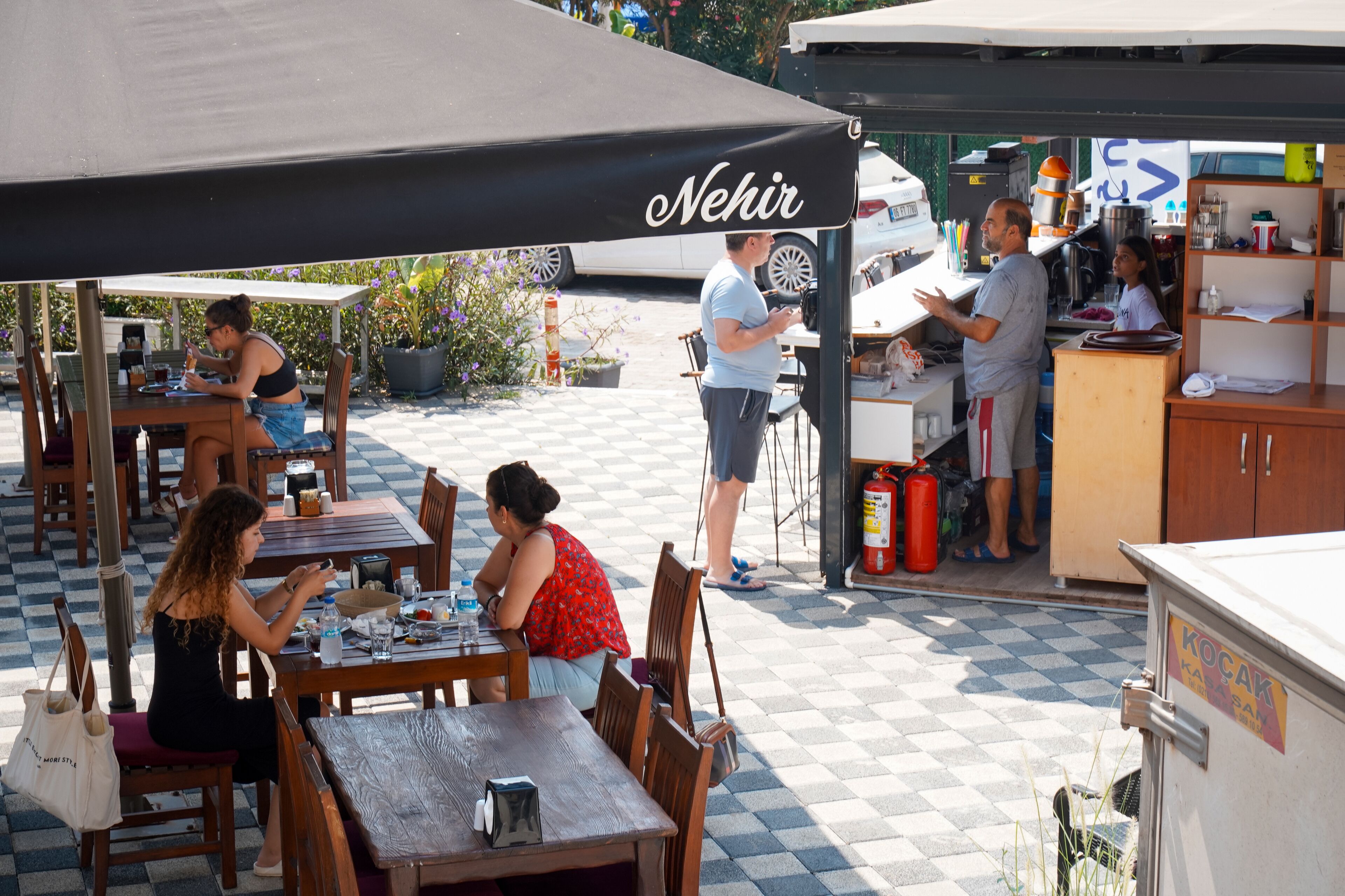 Un café en plein air avec des tables et des chaises en bois, un auvent noir et un espace dédié à la préparation des aliments.