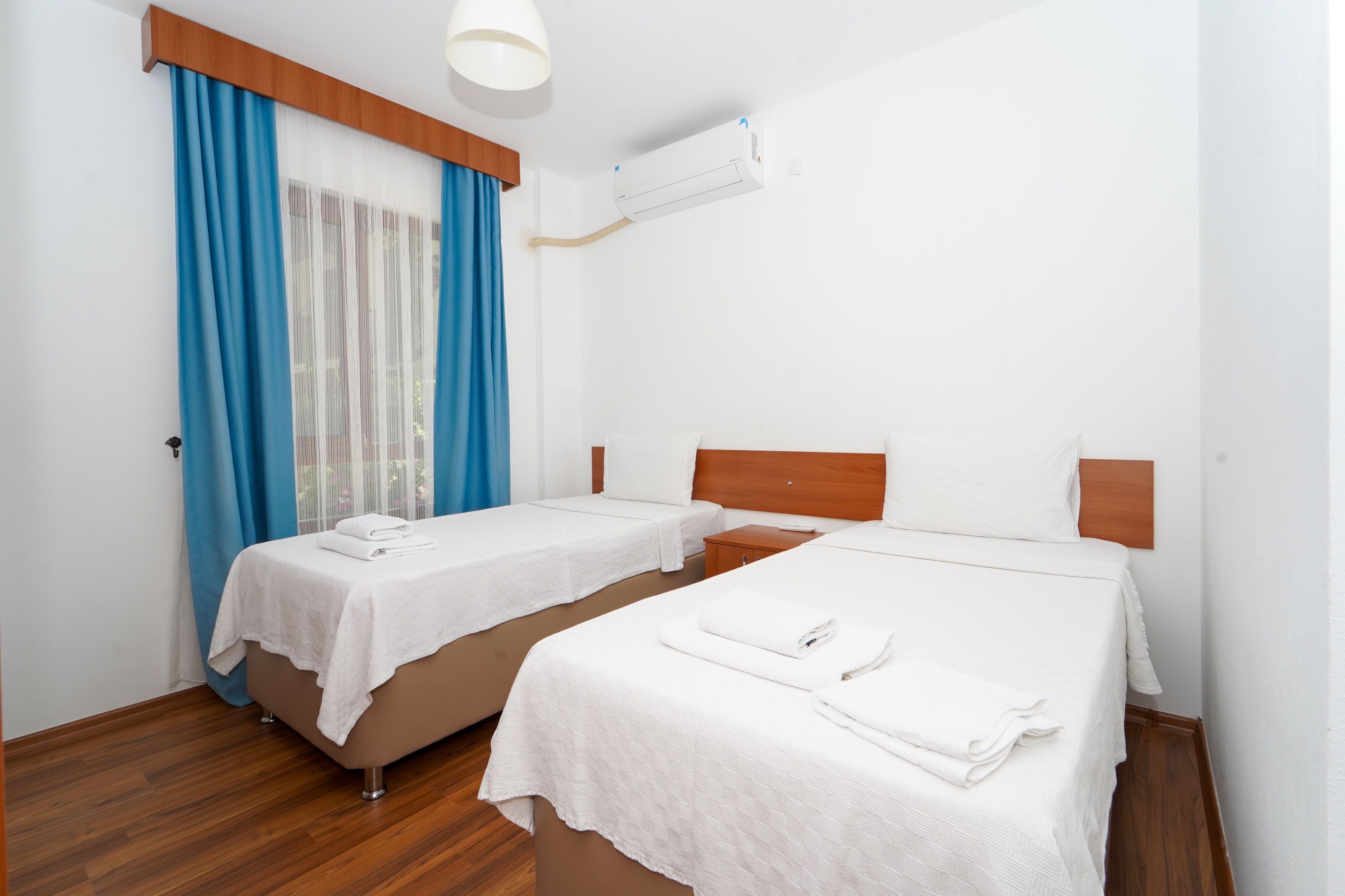 A hotel room with two single beds, a wooden headboard, a window with blue curtains, and an air conditioning unit.