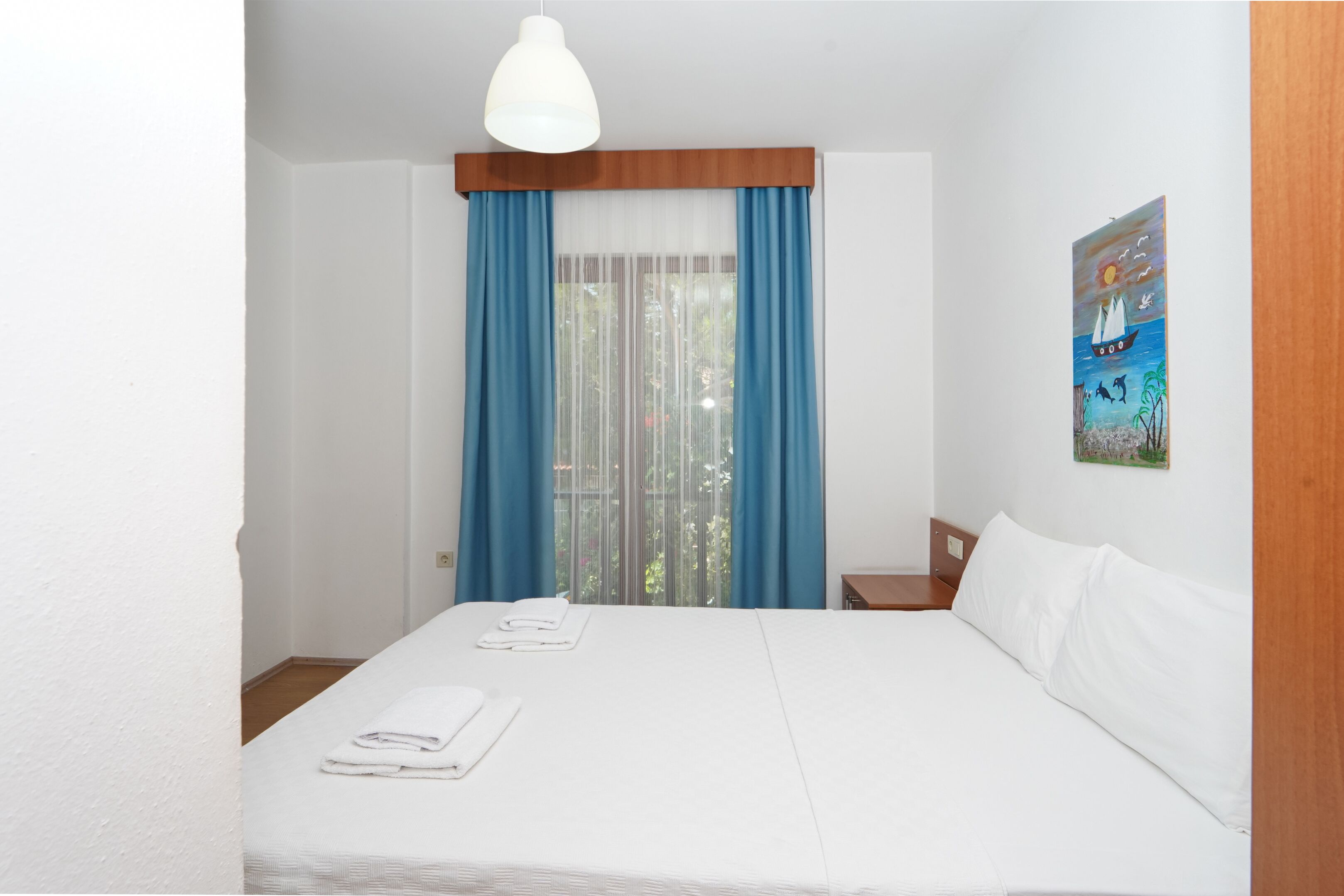 A hotel room with a large bed, white linens, a wooden headboard, a white pendant light, a window with blue curtains, and a painting of a sailboat on the wall.