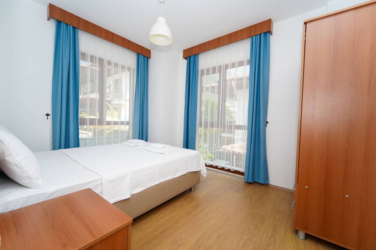 Quarto com cama, cabeceira de madeira, mesa de cabeceira, guarda-roupa e janelas amplas com cortinas azuis.