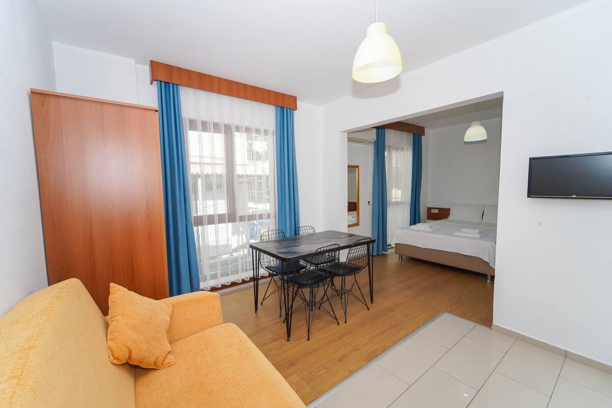 Uma sala de estar moderna com sofá, uma mesa de jantar com cadeiras, uma TV fixada na parede e um quarto separado com cama e banheiro.