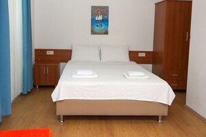 Una cama bien tendida con sábanas blancas y dos toallas dobladas, un armario de madera y un cuadro de un velero en la pared.