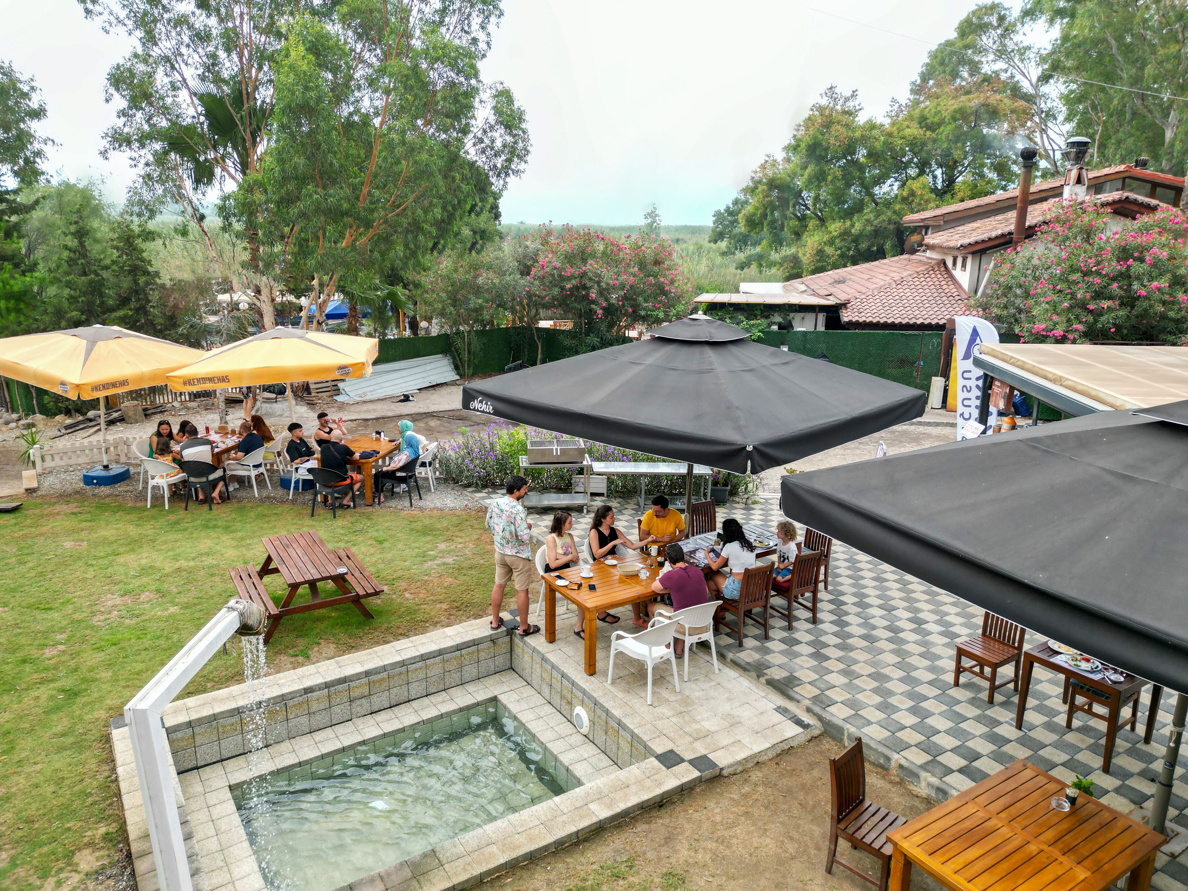 An outdoor patio area with multiple tables and chairs, a pool, and umbrellas providing shade.