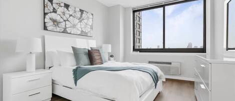 Une chambre moderne avec un grand lit, des tables de chevet blanches, une grande fenêtre donnant sur la ville et un élément décoratif mural.
