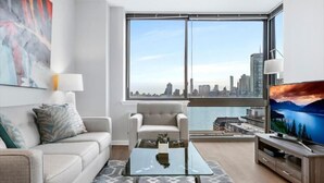 Una sala de estar moderna con un sofá, un sillón, una mesa de centro y un televisor. Un ventanal grande ofrece vistas de la ciudad.