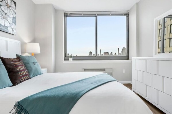 A modern bedroom with a large bed, a window with a city view, a bedside table with a lamp, and a radiator.