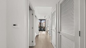 Un couloir étroit aux murs blancs et au plancher en bois mène à une pièce dotée d’une grande fenêtre.