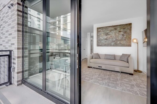 Een moderne woonkamer met een glazen deur naar een balkon, een beige bank, een kamerkleed met patroon en een groot abstract schilderij aan de muur.