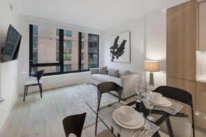 Een moderne woonkamer met een bank, een eettafel voor twee personen, een aan de muur bevestigde televisie en een groot raam met uitzicht op een bakstenen gebouw.