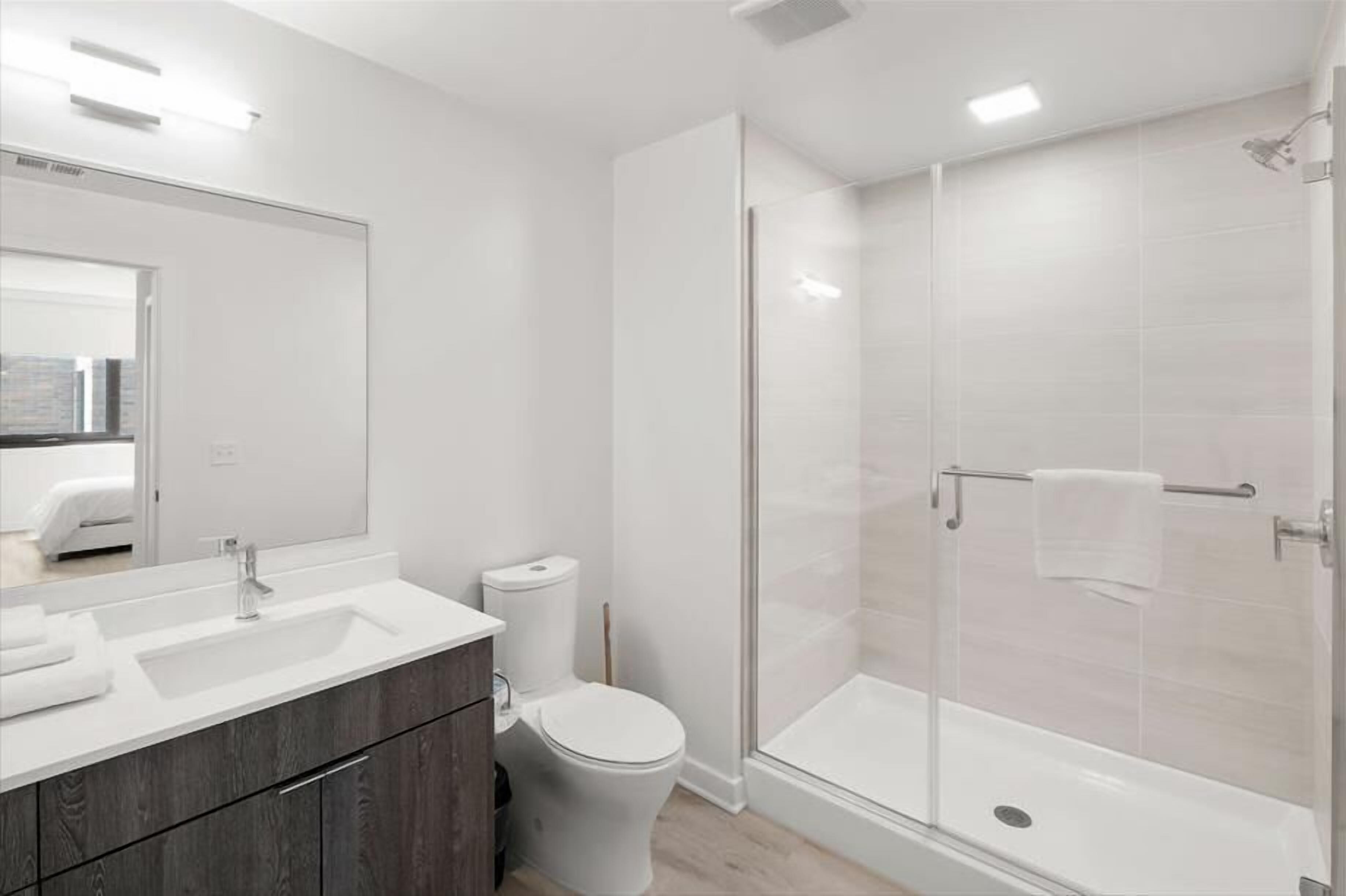 Ein modernes Badezimmer mit einem weißen Waschbecken, einer dunklen Holz-Kommode, einer Toilette und einer gläsernen Duschkabine.