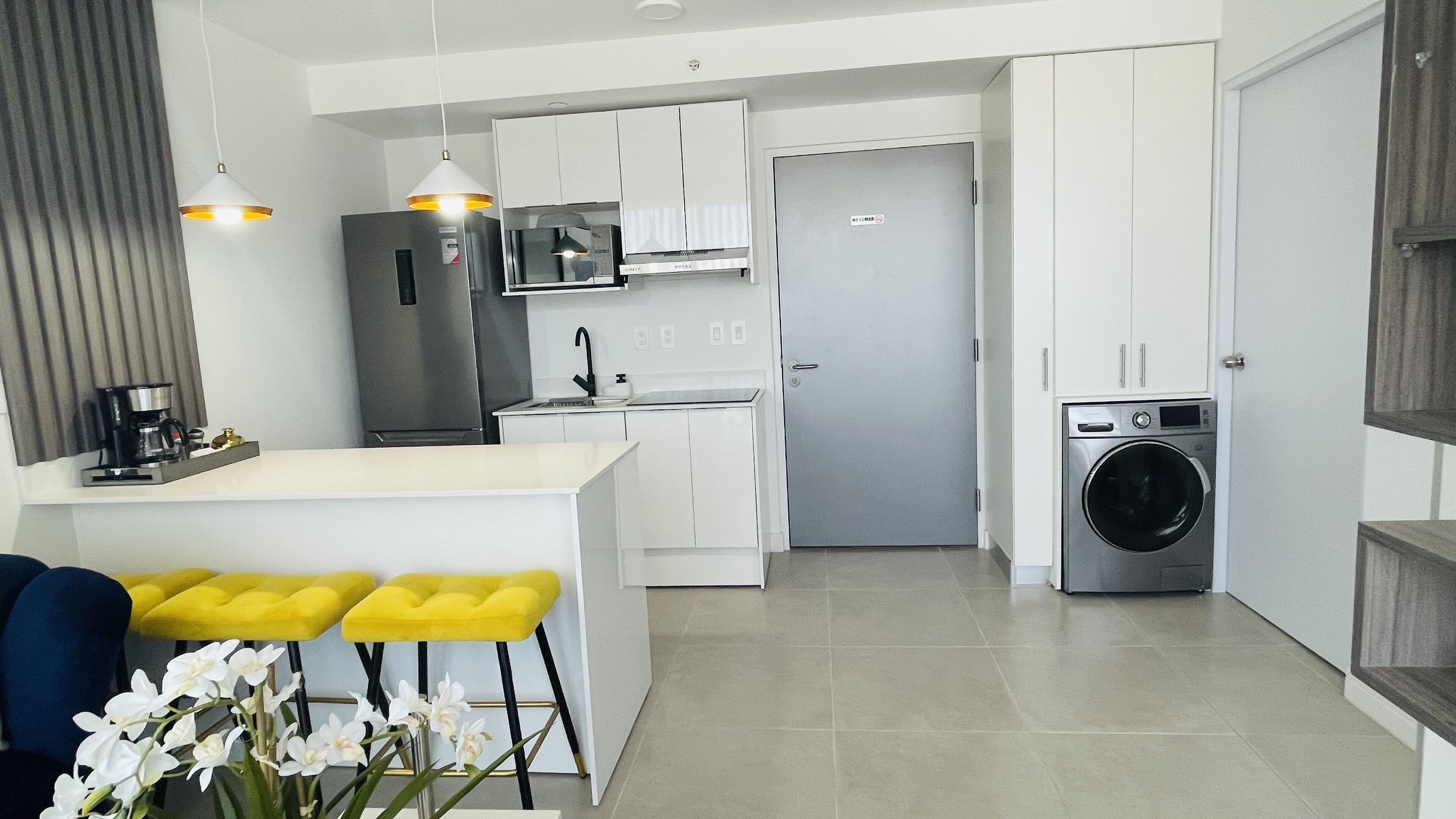 A modern kitchen with white cabinets, a stainless steel refrigerator, a microwave, and a built-in washing machine. There are two yellow bar stools and a coffee machine on the counter.