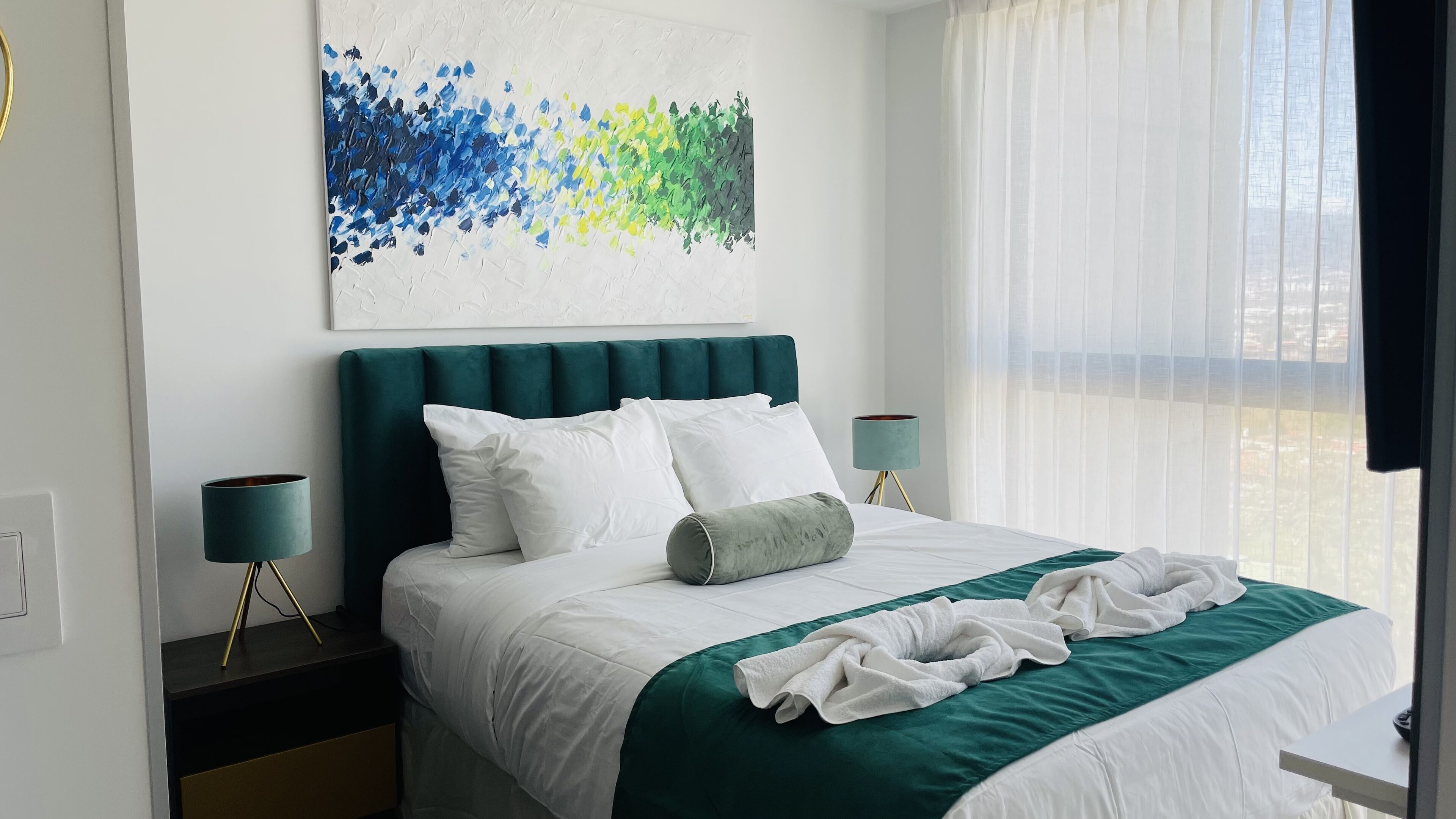 Een opgemaakt bed met wit- en groen beddengoed, een blauwgroen hoofdbord en een modern abstract schilderij erboven.