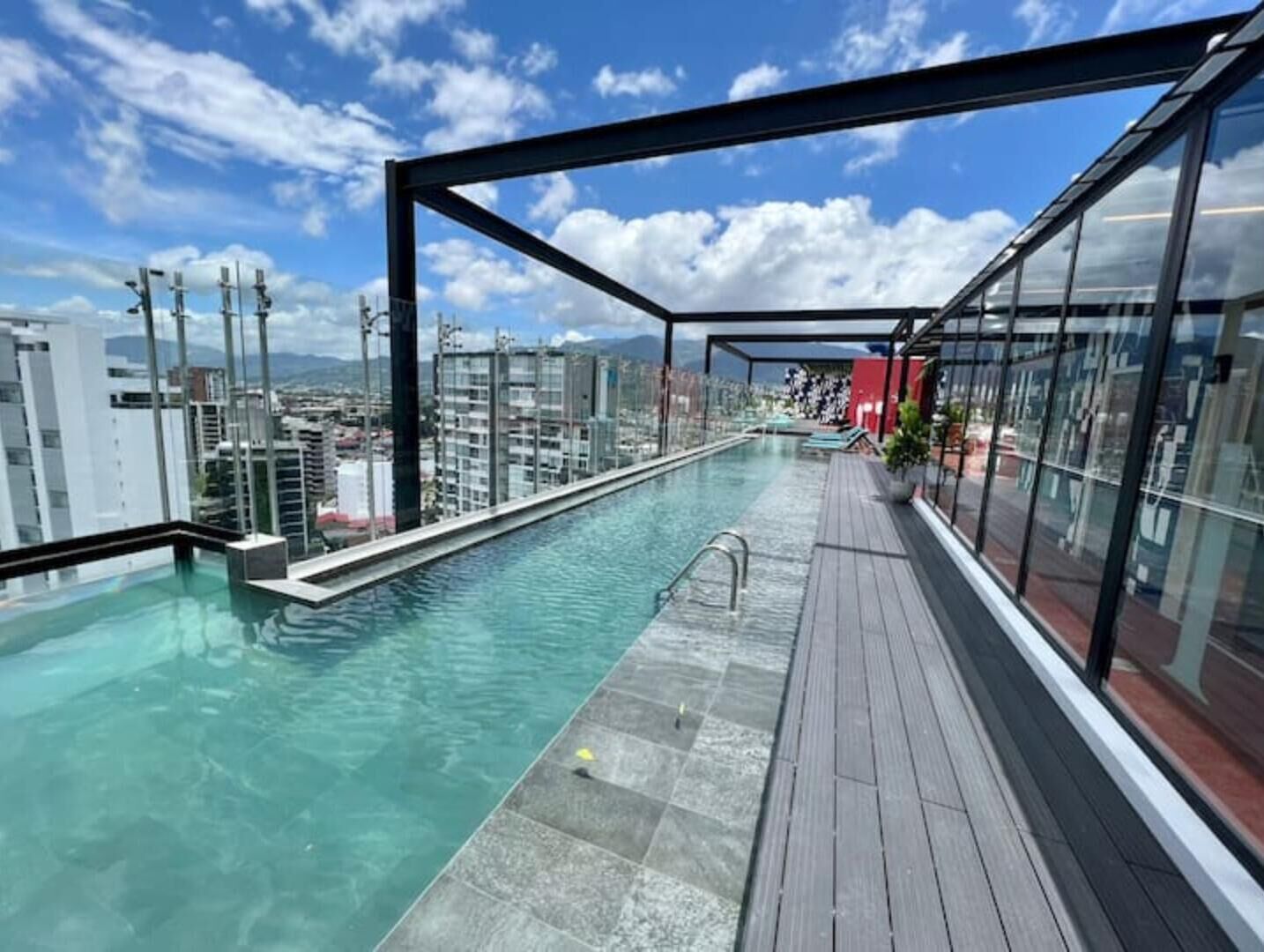 A rooftop infinity pool with a wooden deck and glass walls offering a city view.