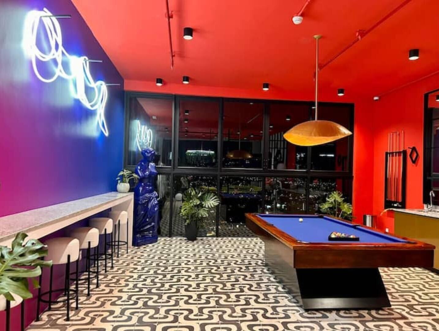 A pool table in a room with a patterned floor, a neon sign, and a bar area with stools.