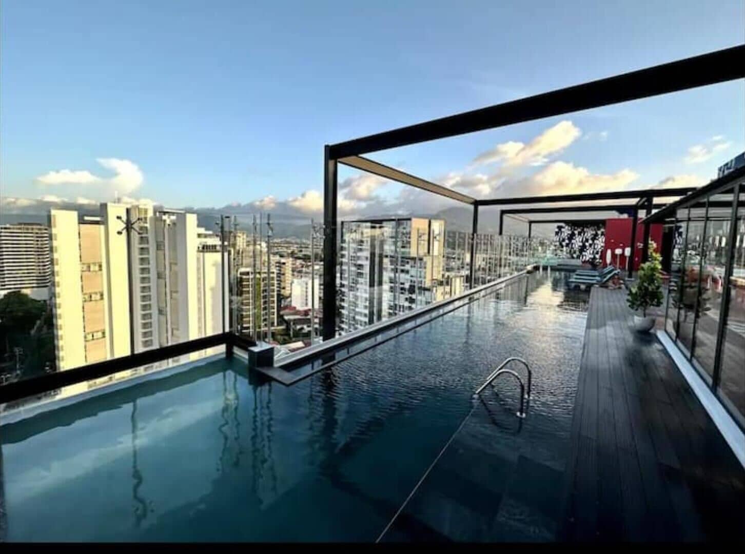 Une piscine à débordement située sur le toit, offrant une vue sur la ville, avec une terrasse en bois et une balustrade en verre.