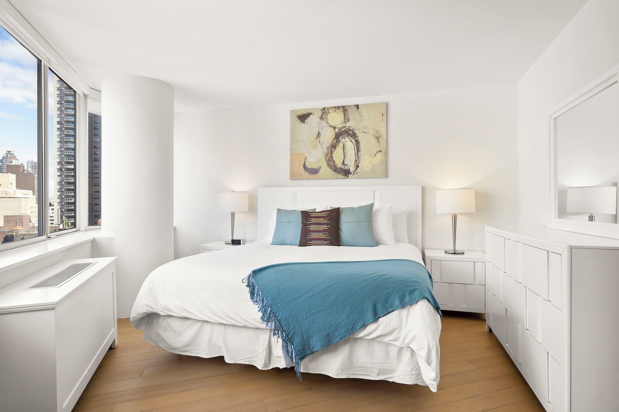 A modern bedroom with a large bed, bedside lamps, a radiator, and a city view.