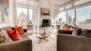 Uma sala de estar moderna com sofá, poltronas, mesa de centro de vidro e televisão. Grandes janelas proporcionam vista para a cidade.
