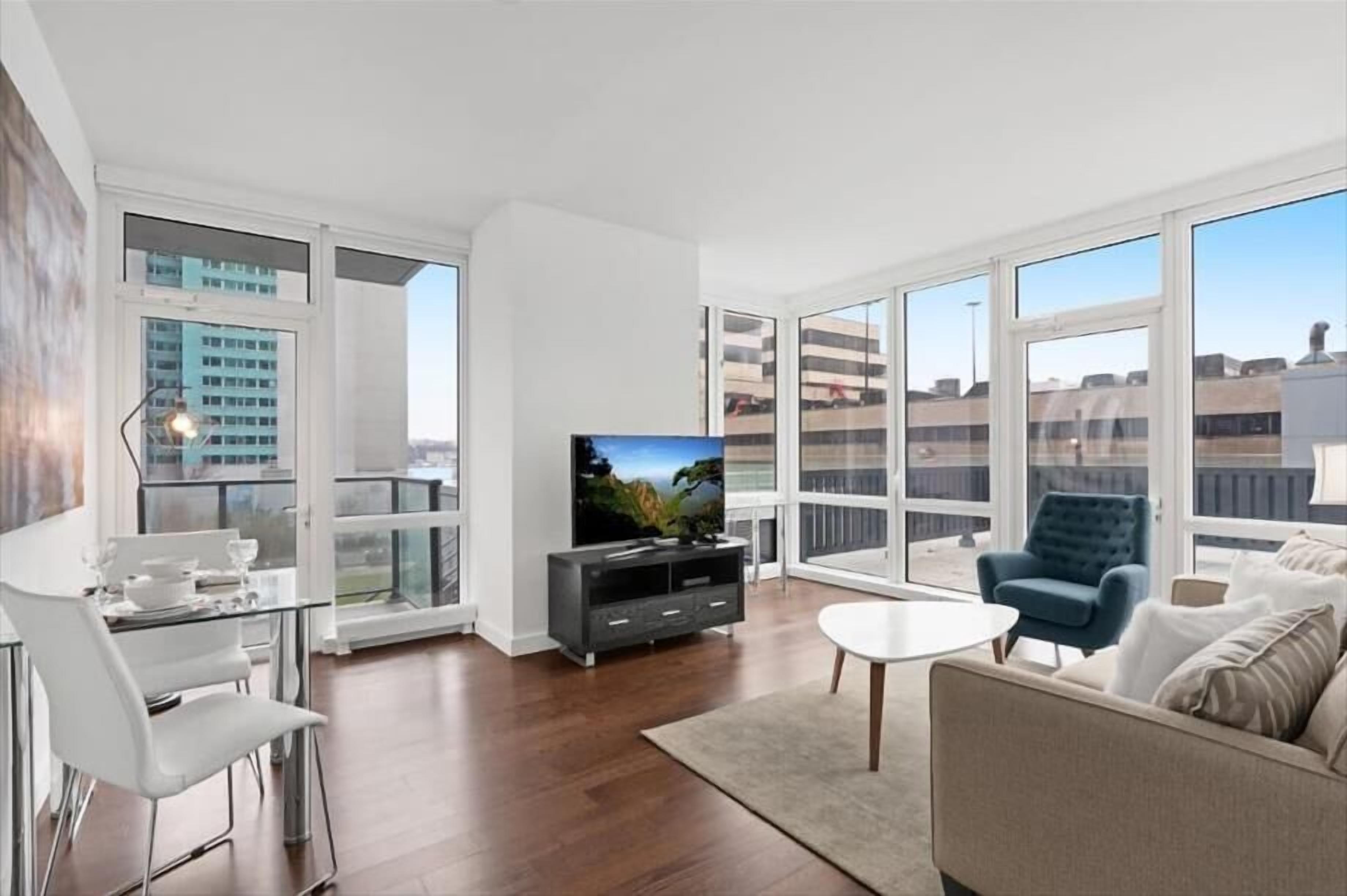 Een moderne woonkamer met een flatscreen televisie, een eethoek met een glazen tafel en stoelen, en grote ramen met uitzicht op de stad.