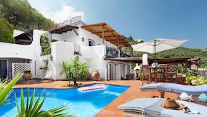 A pool area with a white building, outdoor seating, and a view of the hills. - YourHome - Villa Mia (Massa Lubrense)